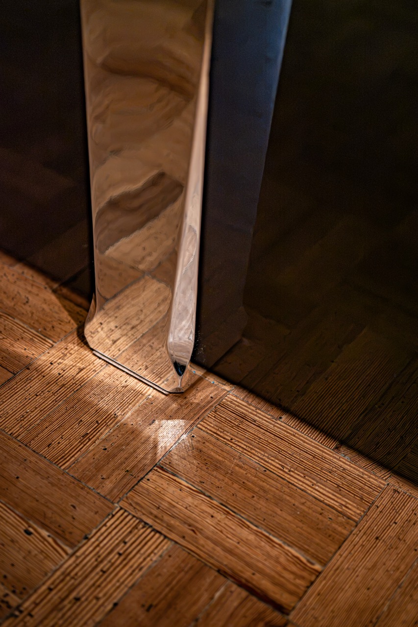 A close-up of a shiny metal table leg reflecting light, standing on a wooden parquet floor with a geometric pattern.
