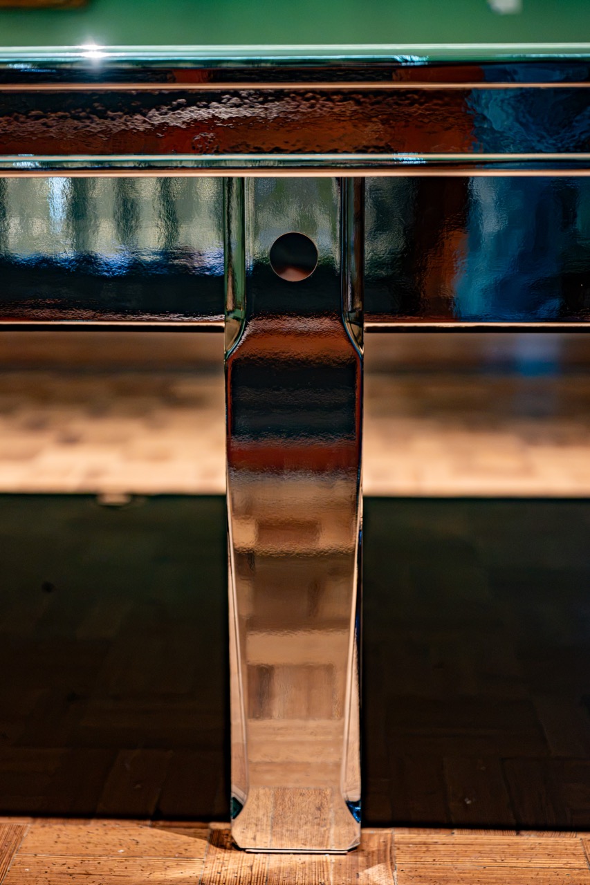 A close-up of a metallic, reflective table leg on a wooden floor, with a circular hole near the top of the leg and blurred colours in the background.