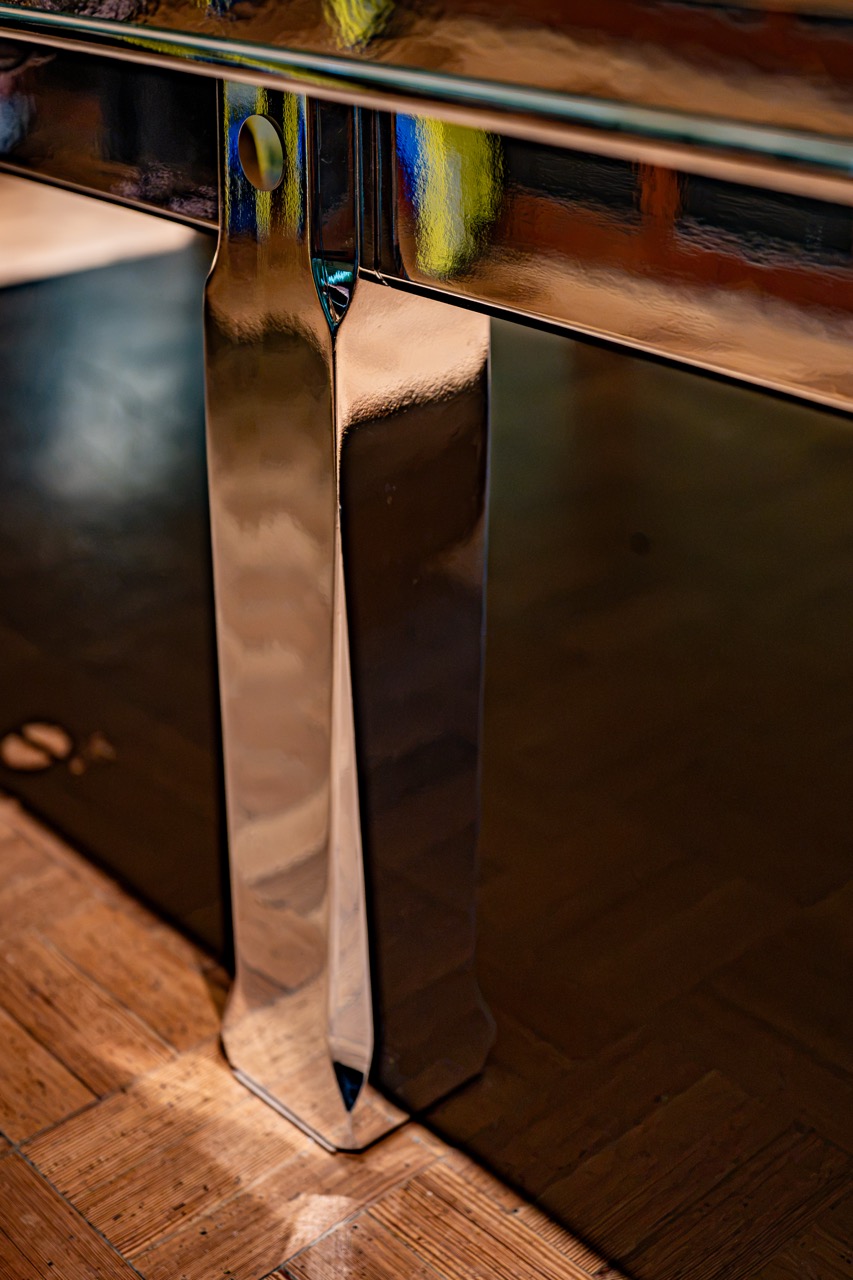 Close-up of a shiny metal table leg with reflections, standing on a wooden parquet floor.