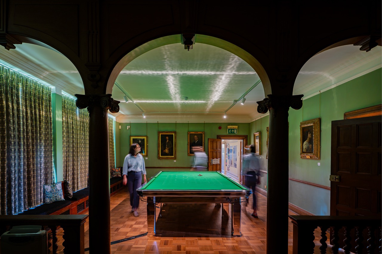 A green billiards table sits in a classic room with wooden floors and portraits on the walls. Two blurred figures walk near the table, suggesting motion. The scene is viewed through an arched doorway.