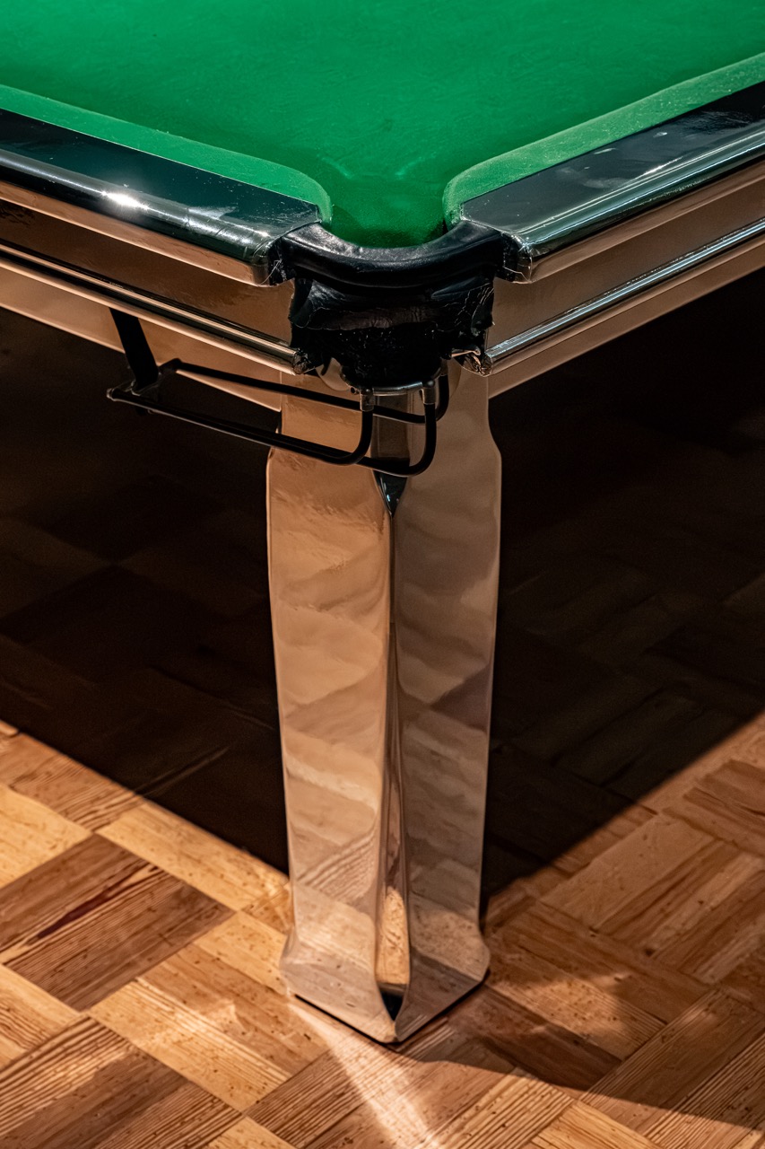 A close-up of the corner of a shiny metal pool table with a green baize surface, showing the pocket and reflecting parquet wood flooring below.