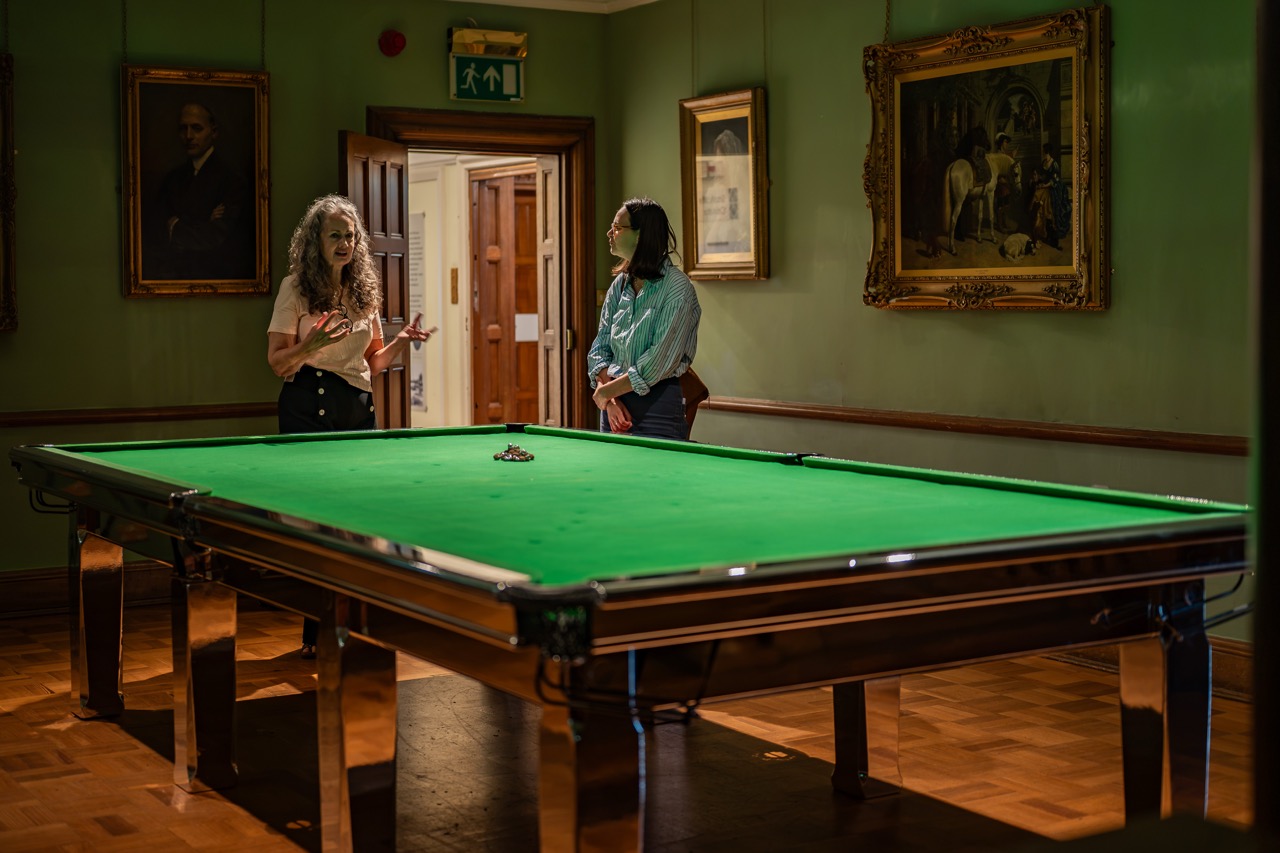 Two women stand and talk beside a green snooker table in a warmly lit, art-filled room with wooden floors and classic framed paintings on the walls.