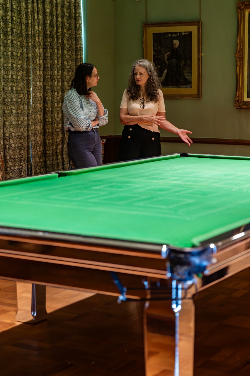 Two women stand and talk beside a green snooker table in a room with framed portraits on the walls and a draped window curtain in the background.