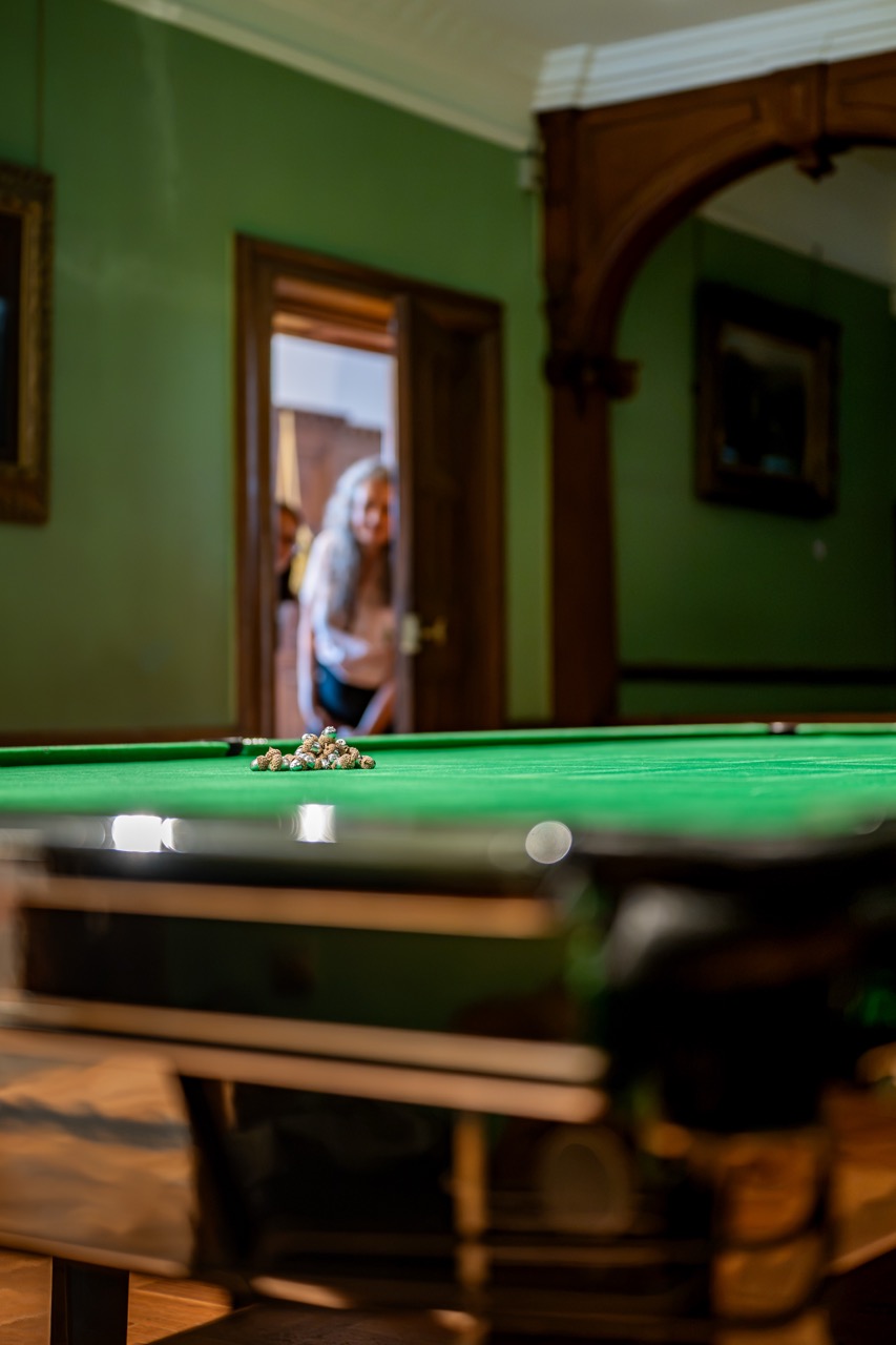 A woman stands in the doorway of a room with green walls, looking towards a snooker table with balls grouped in the centre. The image foreground is blurred, focussing on the door and woman.