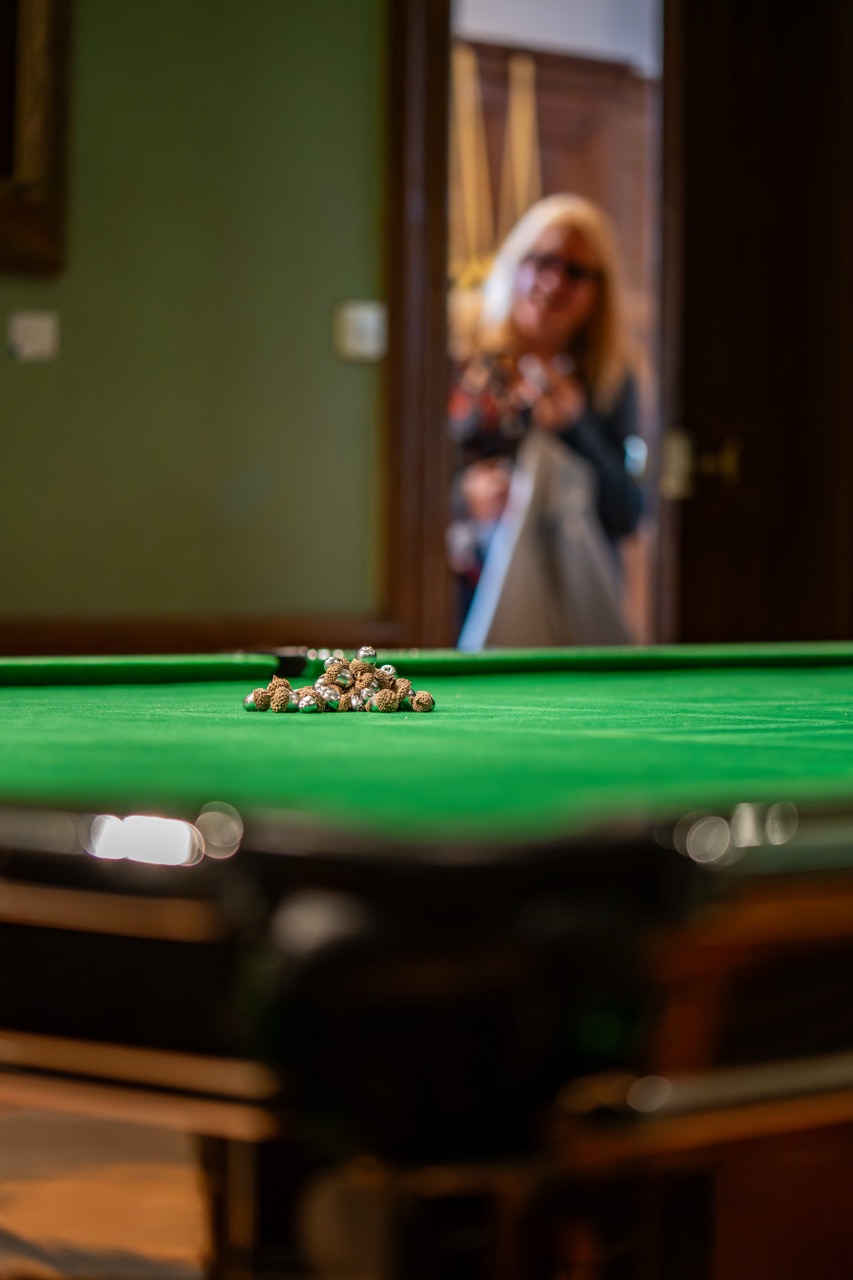 A blurred person stands in the background while a snooker or pool table with a small pile of nuts rests in focus on the green baize surface in the foreground.