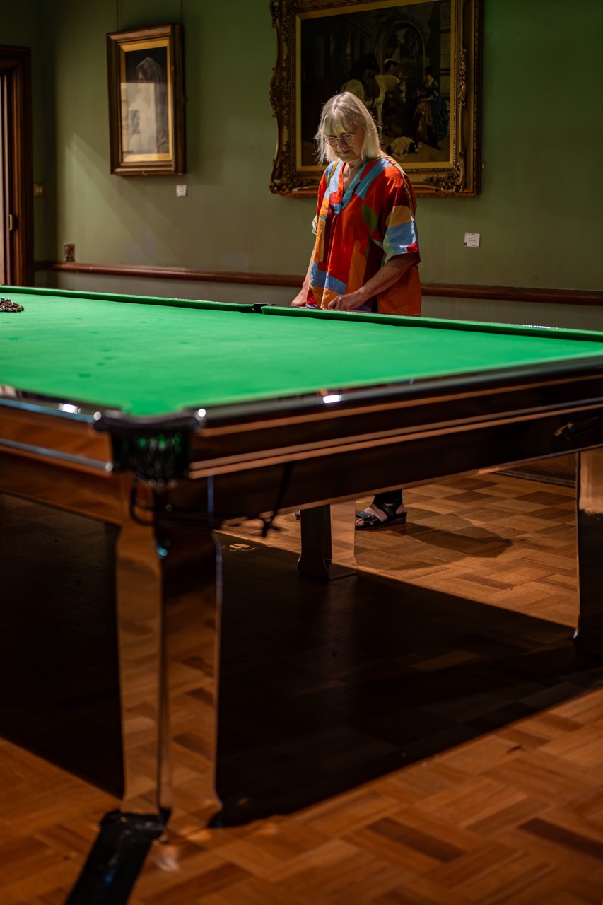 A person with long blond hair wearing a colourful shirt stands by a snooker table in a room with wooden floors and framed paintings on green walls.