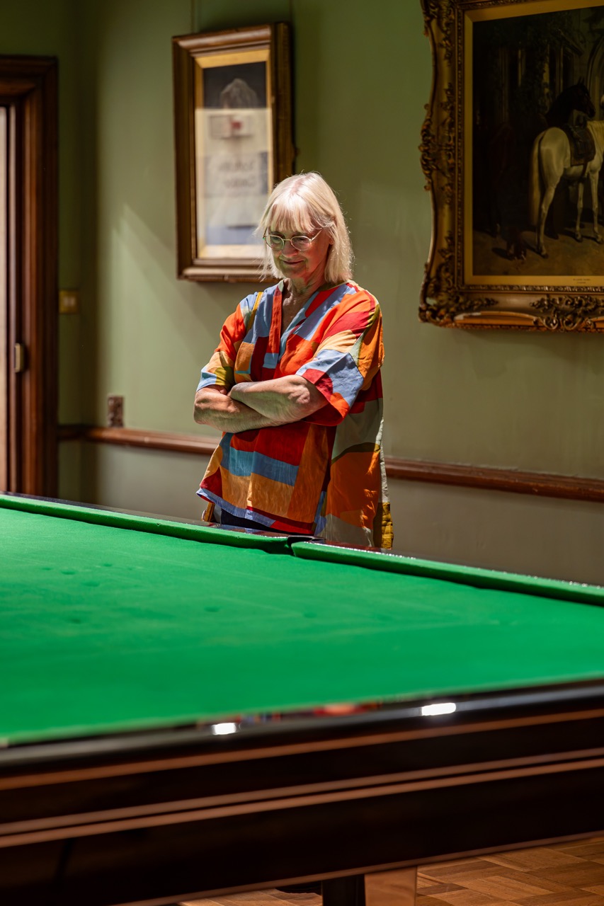 An older woman with long white hair and glasses stands with folded arms beside a green snooker table, wearing a colourful shirt, in a room decorated with framed paintings and certificates.