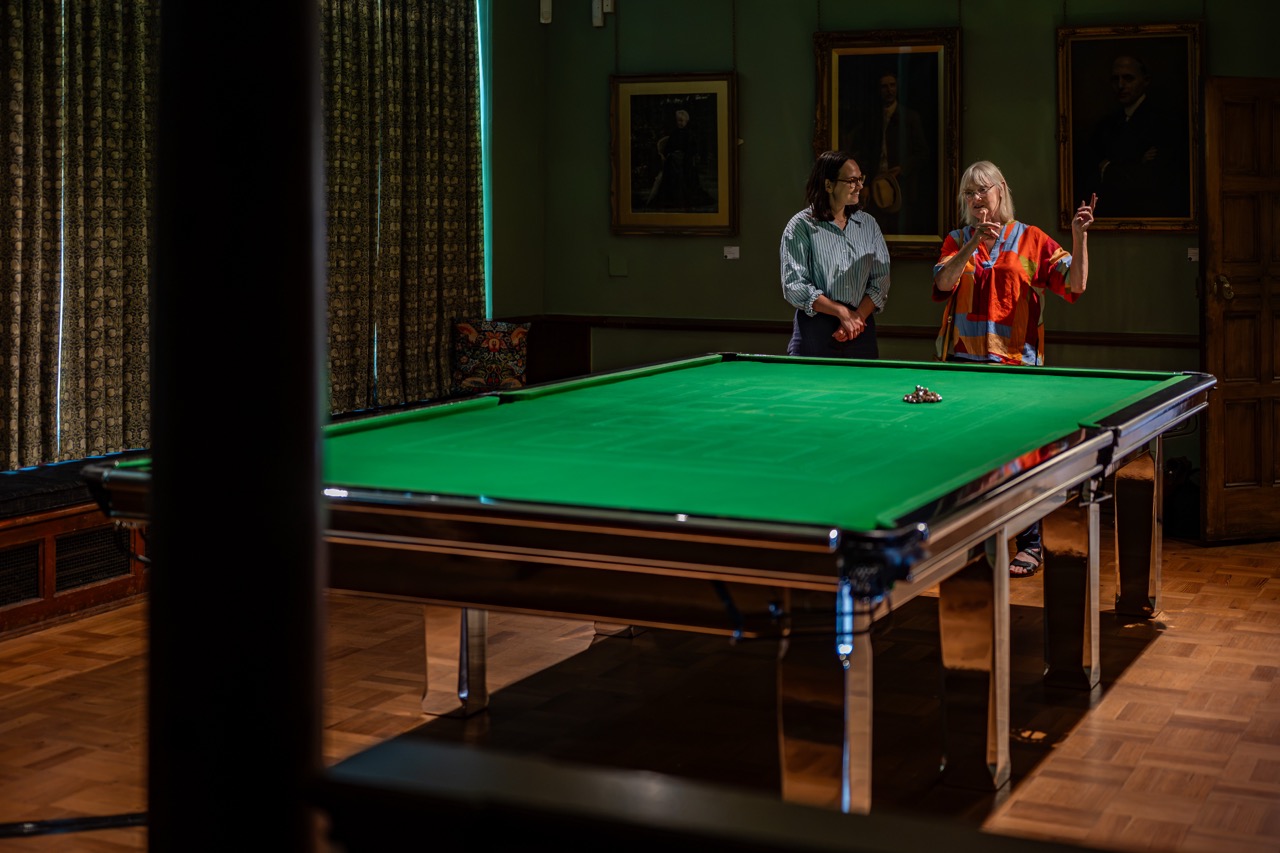 Two women stand and talk beside a green snooker table in a room with wooden floors, framed portraits on the wall, and closed patterned curtains.