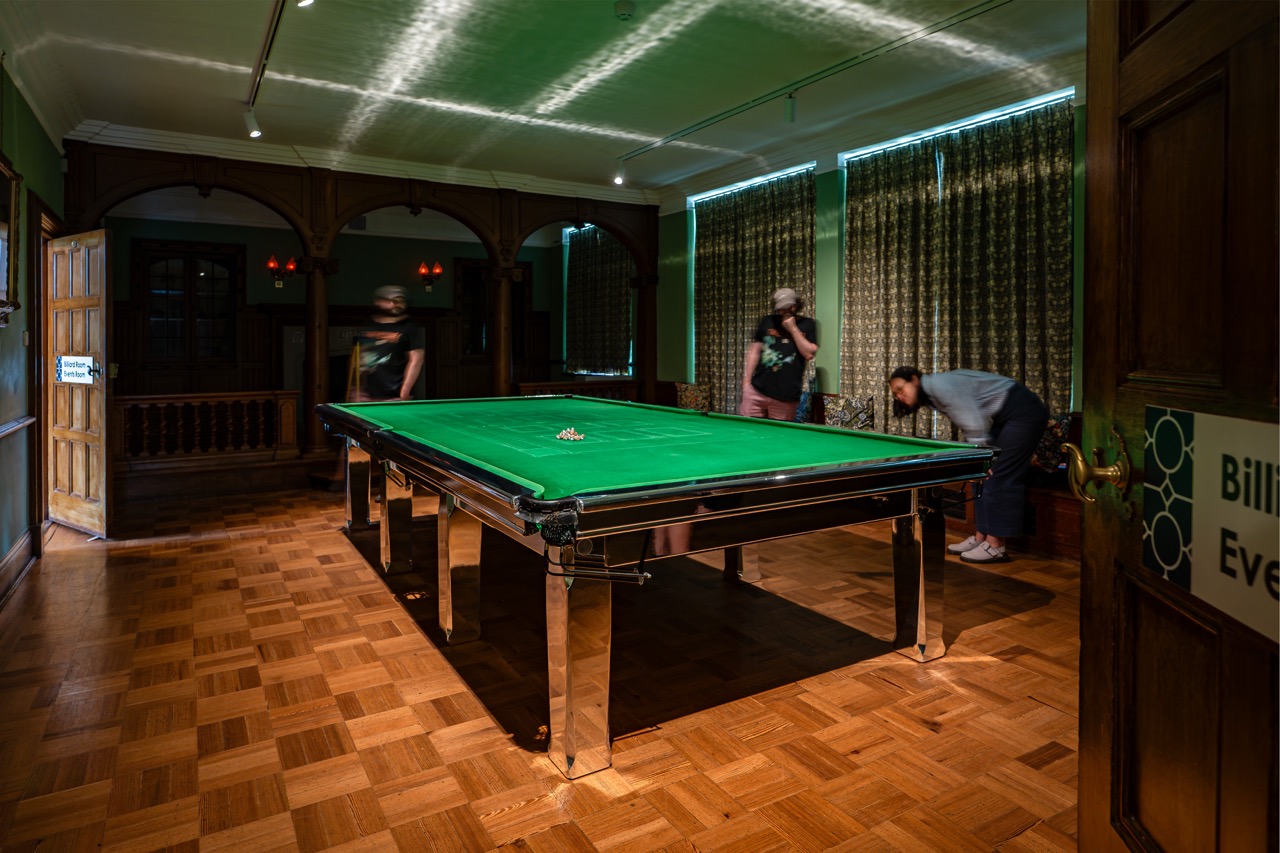 Three people set up to play billiards in a wood-panelled room with parquet flooring, curtains, and a green billiard table under ceiling lights.
