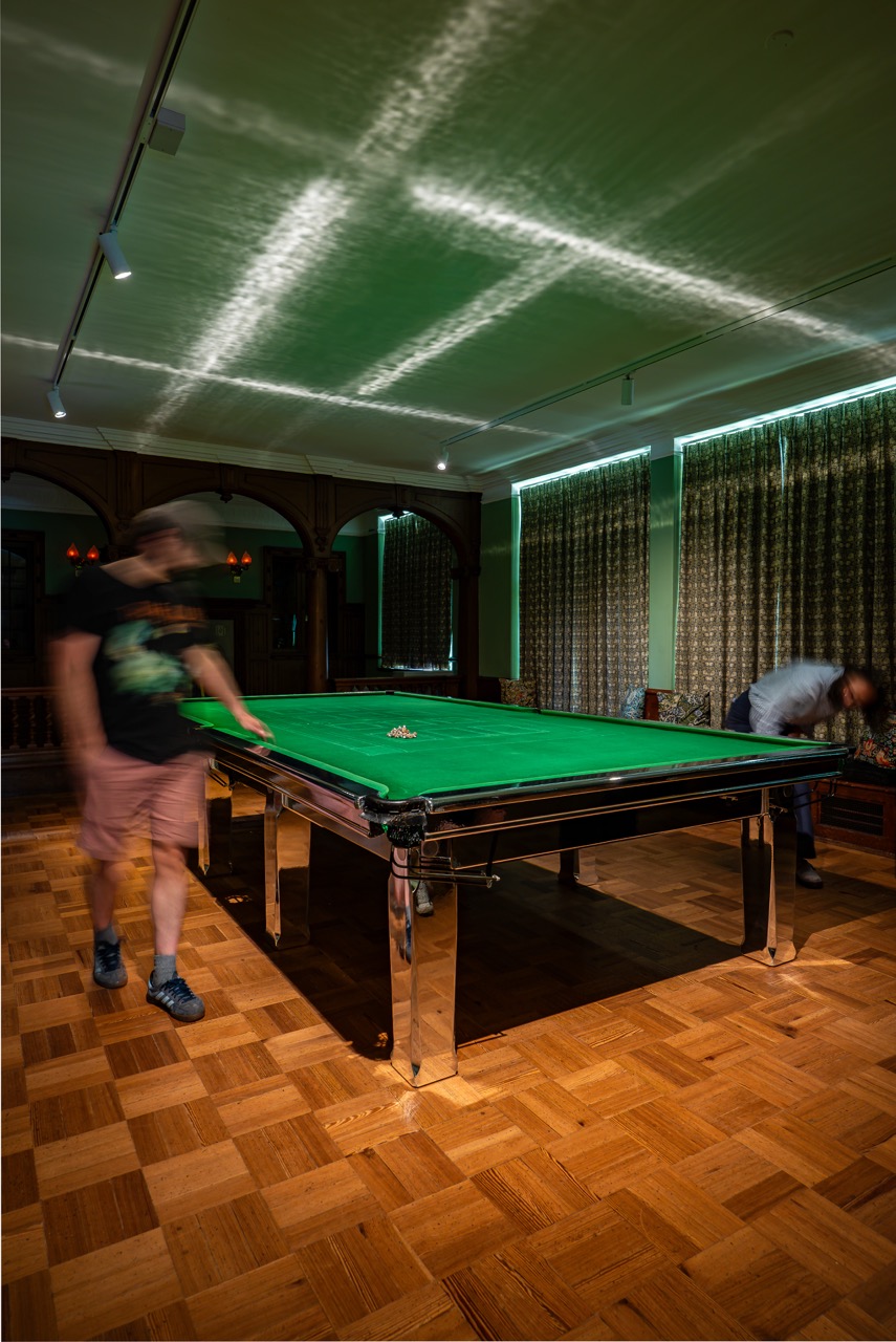 Two people play snooker in a room with patterned curtains and wooden floors. Light patterns are reflected on the ceiling above the green snooker table. Both people appear blurred from motion.