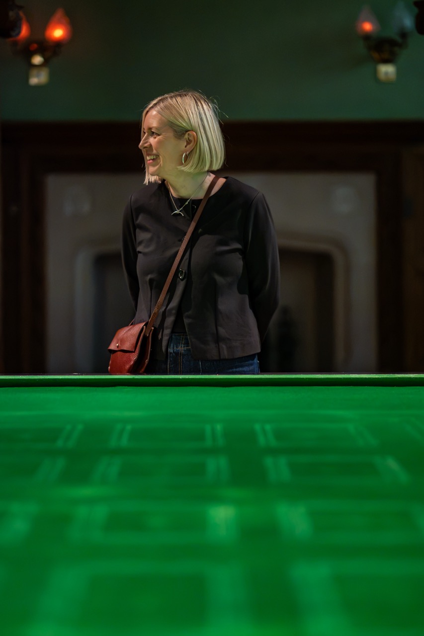 A woman with blonde hair, wearing a dark top and crossbody bag, stands and smiles, looking to her right. She is behind a large table with a green, patterned surface in a warmly lit room.