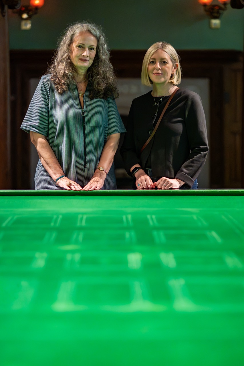 Two women stand side by side behind a brightly lit green snooker or pool table, looking at the camera in a warmly lit indoor setting.