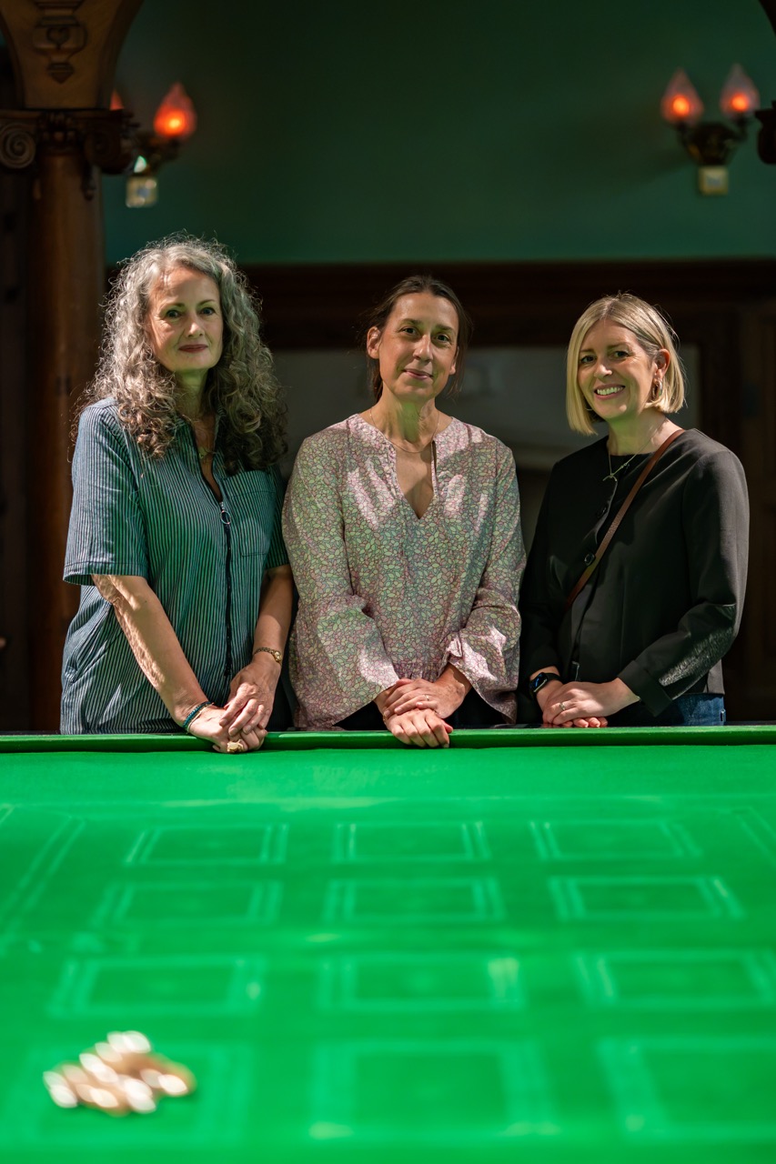 Three women stand side by side, smiling, behind a green billiards or snooker table with a small pile of balls on it. The room is dimly lit with ornate wooden columns and wall-mounted lights in the background.