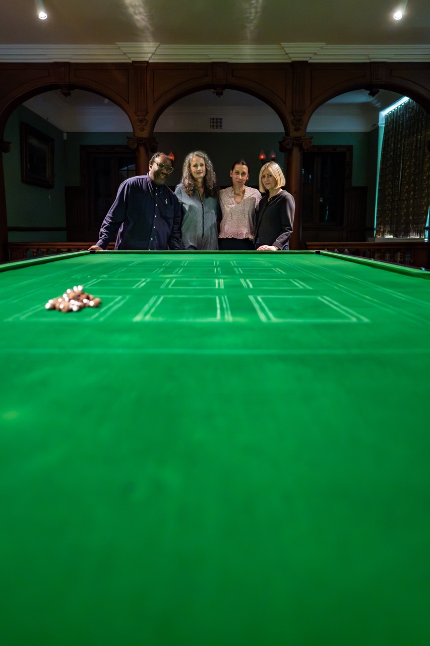 Four people stand together at the end of a large, green gaming table in a dimly lit room, with a pile of chips placed on the table in the foreground.