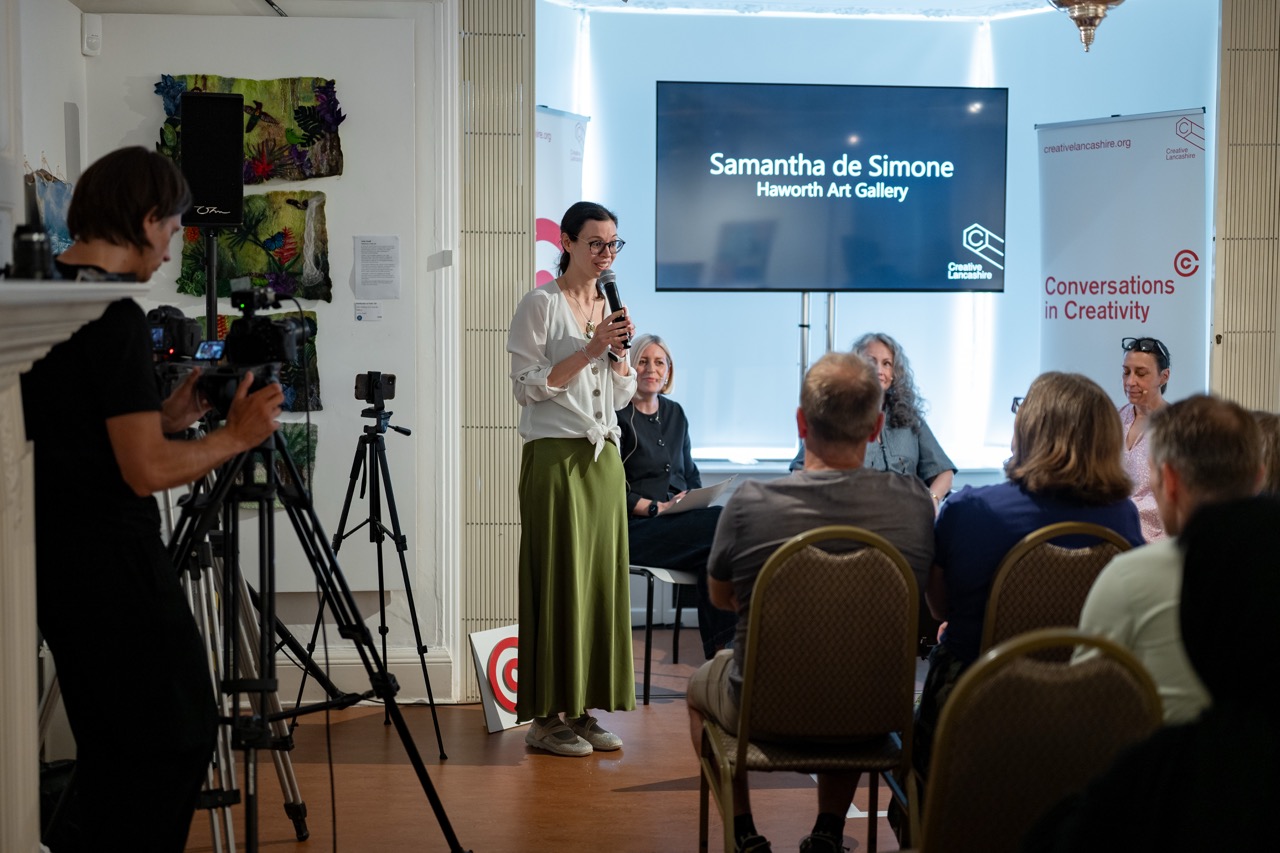 A woman stands and speaks into a microphone in front of a seated audience. Behind her, a screen reads Samantha de Simone, Haworth Art Gallery. A camera operator films the event. Four panellists sit on stage.