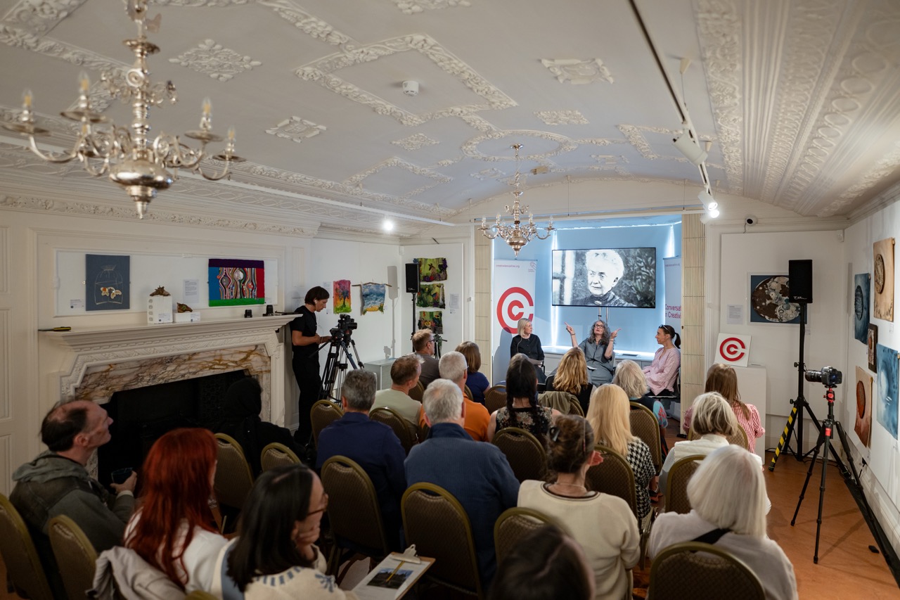 An audience watches a panel discussion in a decorated room with art on the walls. Two cameras record the event, and three panellists sit at the front under a window displaying artwork. A chandelier hangs from the ornate ceiling.