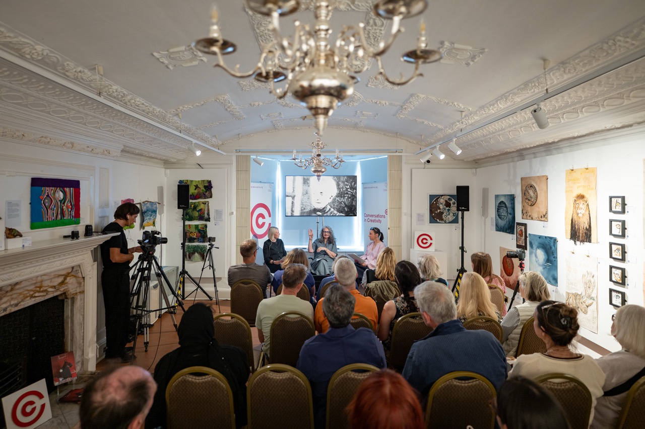 A seated audience watches a panel of three speakers in an art gallery. A camera operator films the event. Colourful artwork decorates the walls, and a chandelier hangs from the ornate ceiling.