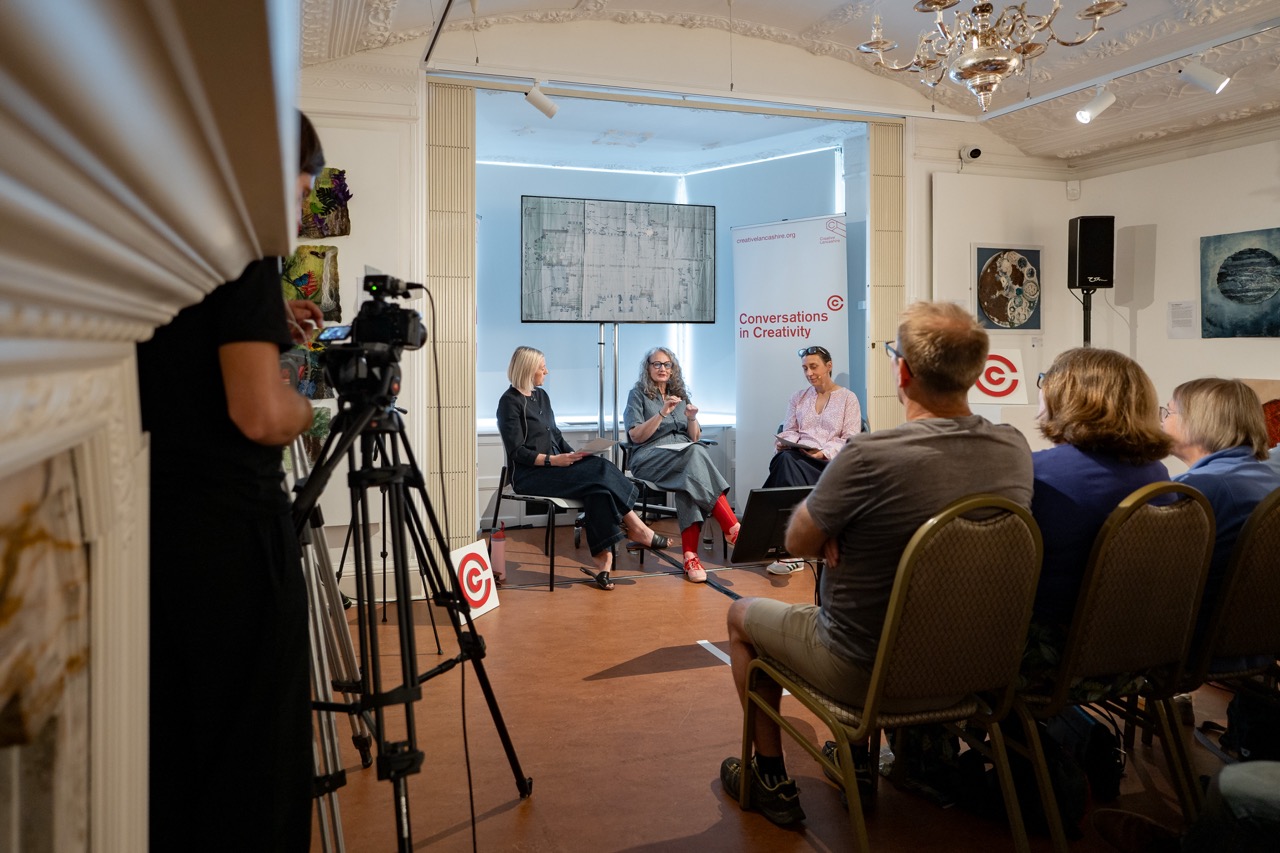 Two women and one man have a panel discussion in front of an audience in a gallery. A camera operator films the event. The backdrop reads Conversations in Creativity and art hangs on the walls.