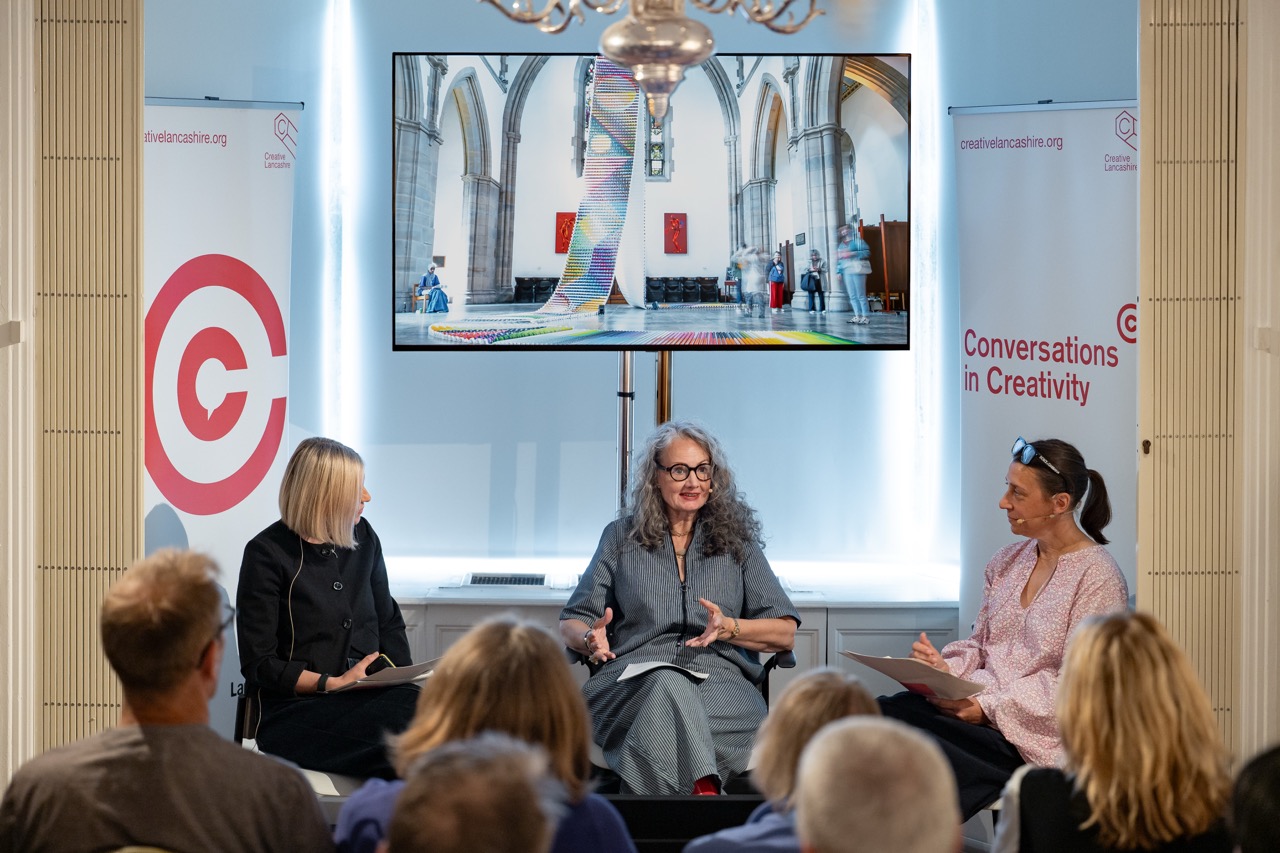 Three women sit and talk onstage in front of an audience, with a large screen behind them showing art inside a building. Banners read Conversations in Creativity.