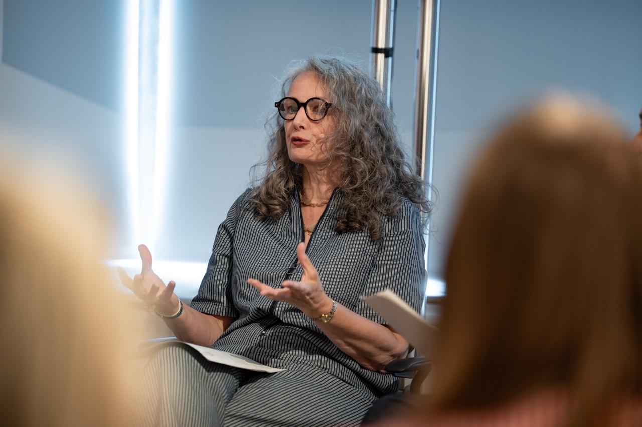 A woman with long grey hair and glasses speaks while seated, gesturing with her hands, in a well-lit room. She wears a striped outfit and holds papers. Blurred figures are visible in the foreground.
