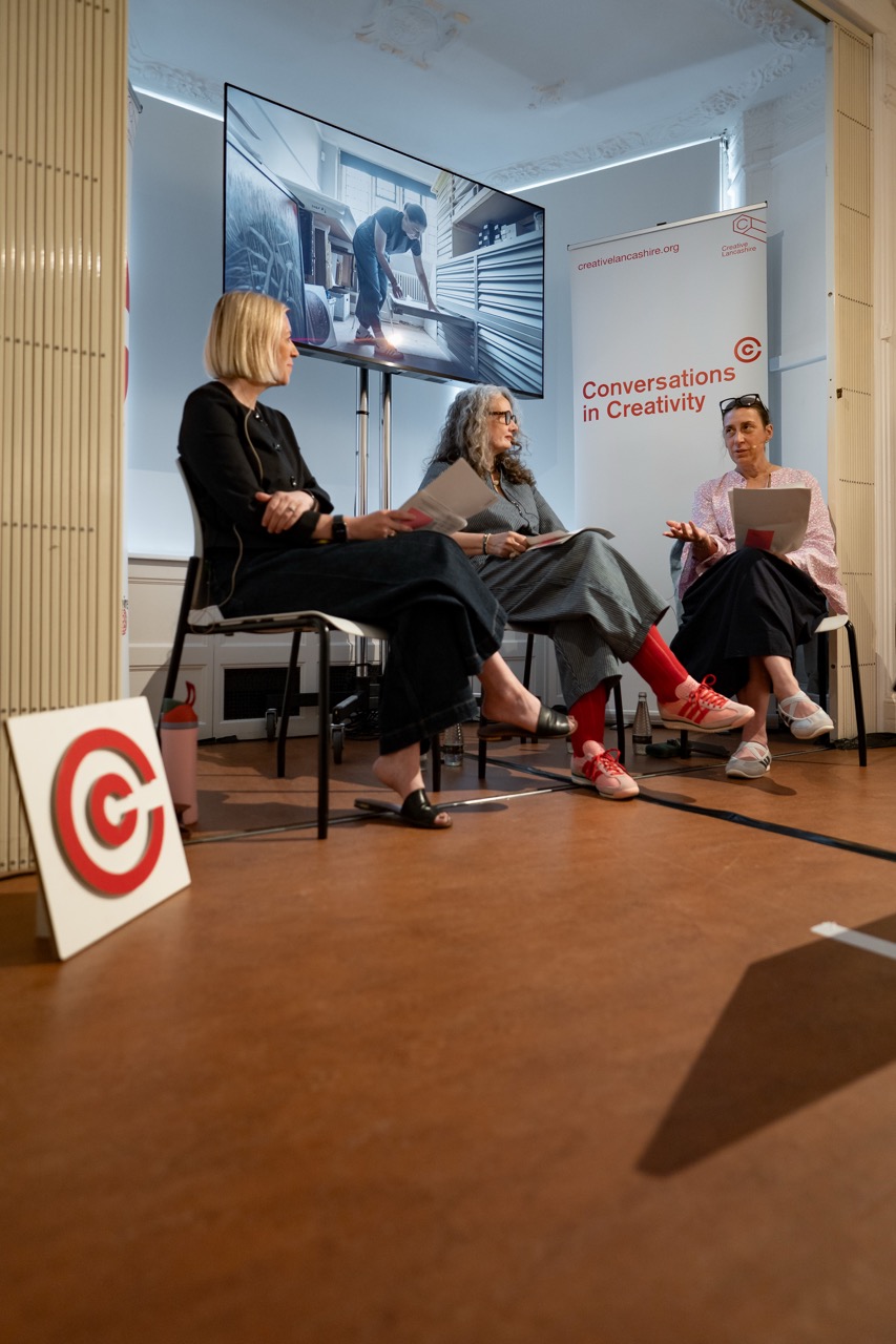 Three women sit in chairs, engaged in discussion. Behind them is a large screen displaying a man working, and a banner reading Conversations in Creativity. A sign with a red C is on the floor.