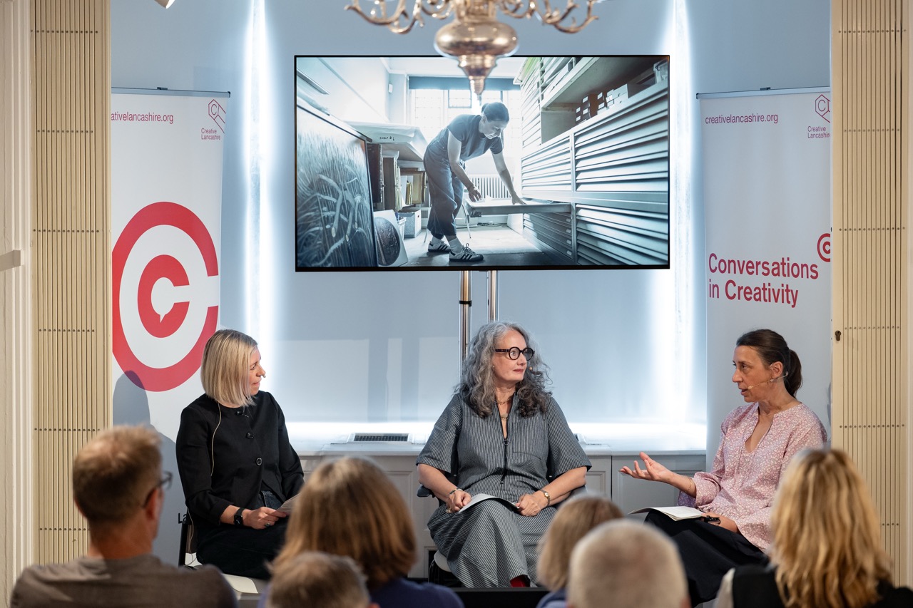 Three women sit and talk on stage at a Conversations in Creativity event, with a screen behind them showing a person working with metal in a workshop. Audience members watch the discussion.