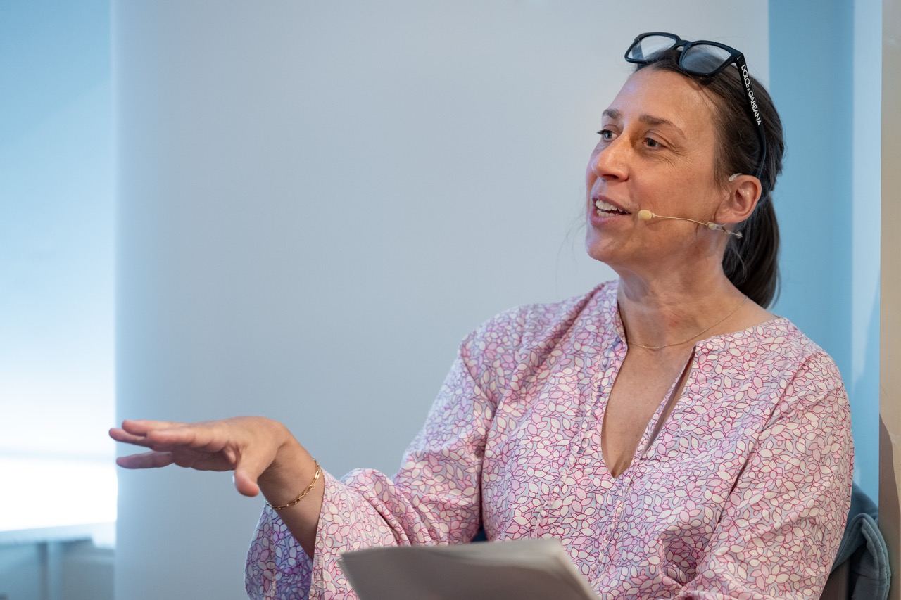 A woman wearing glasses on her head and a light-patterned blouse speaks into a headset microphone while gesturing with her hand. She holds papers in her lap and sits against a light-coloured wall.