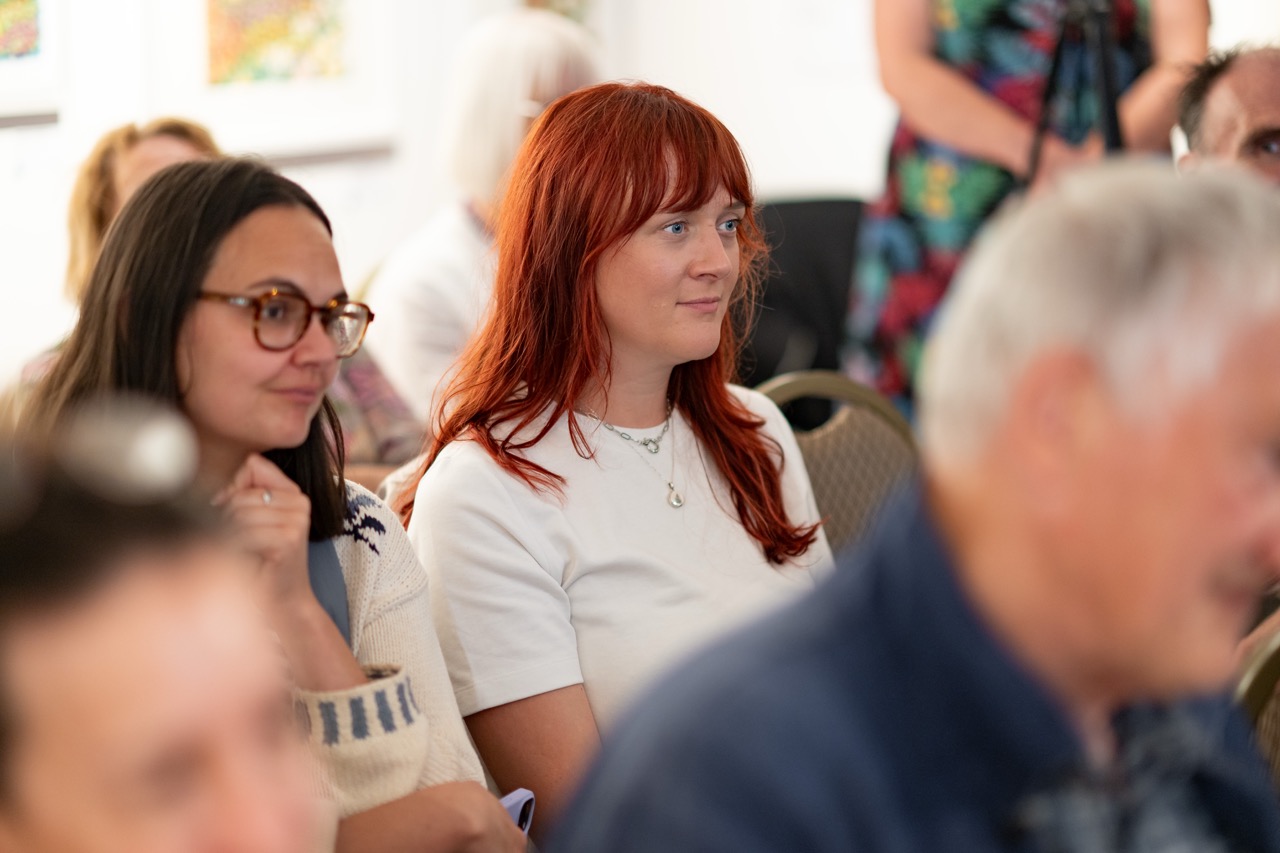 A group of people sit closely together at an indoor event. A woman with long red hair and a white shirt is in focus, sitting beside a woman with glasses and dark hair. Other attendees are visible in the blurred background.
