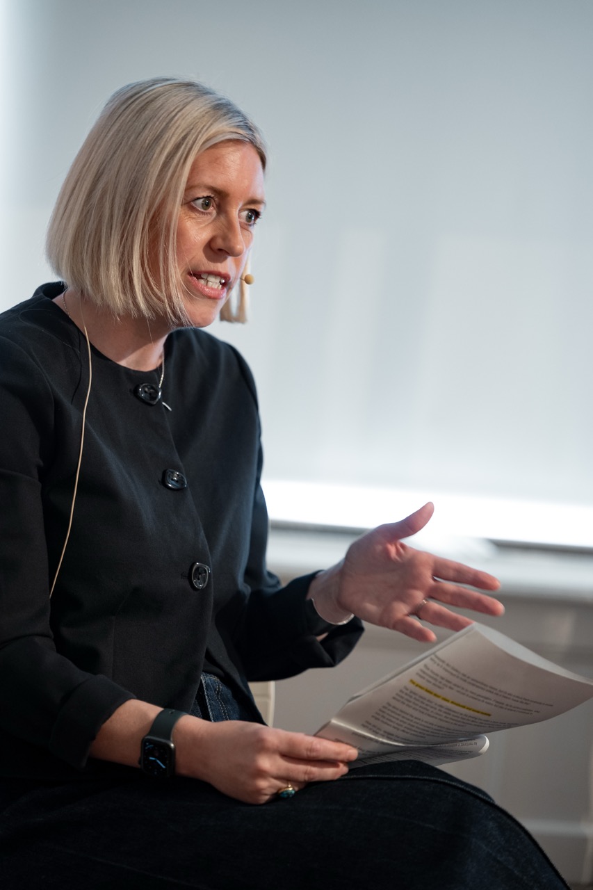 A woman with short blonde hair speaks whilst holding papers. She wears a black jacket and a headset microphone, gesturing with her left hand. The background is softly lit and out of focus.