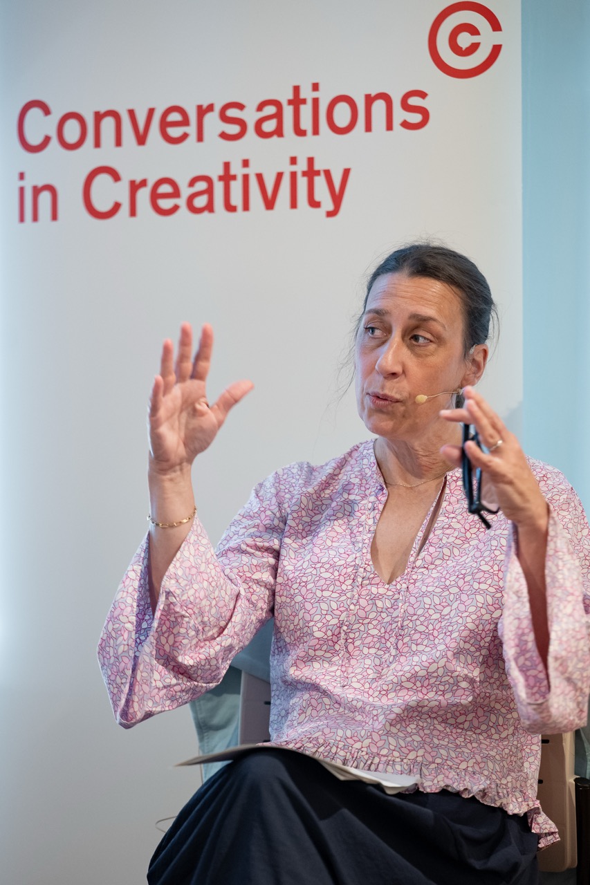A woman speaks into a headset microphone, gesturing with one hand and holding spectacles in the other, seated in front of a sign that reads Conversations in Creativity..