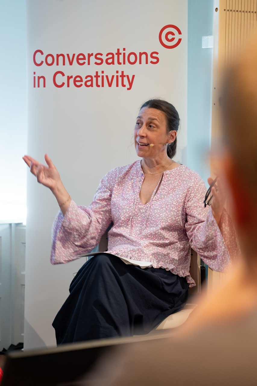 A woman in a pink floral blouse gestures whilst speaking during an event, seated in front of a banner that reads Conversations in Creativity.