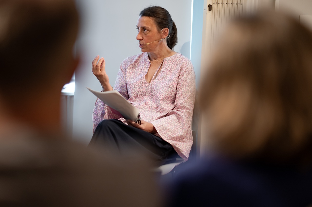 A woman with a headset and floral blouse speaks whilst holding papers, seated indoors. Two blurred figures are visible in the foreground, facing her.