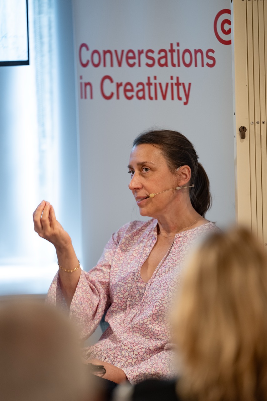 A woman wearing a headset sits and gestures whilst speaking at an event. Behind her is a sign that reads Conversations in Creativity. Audience members are visible in the foreground.