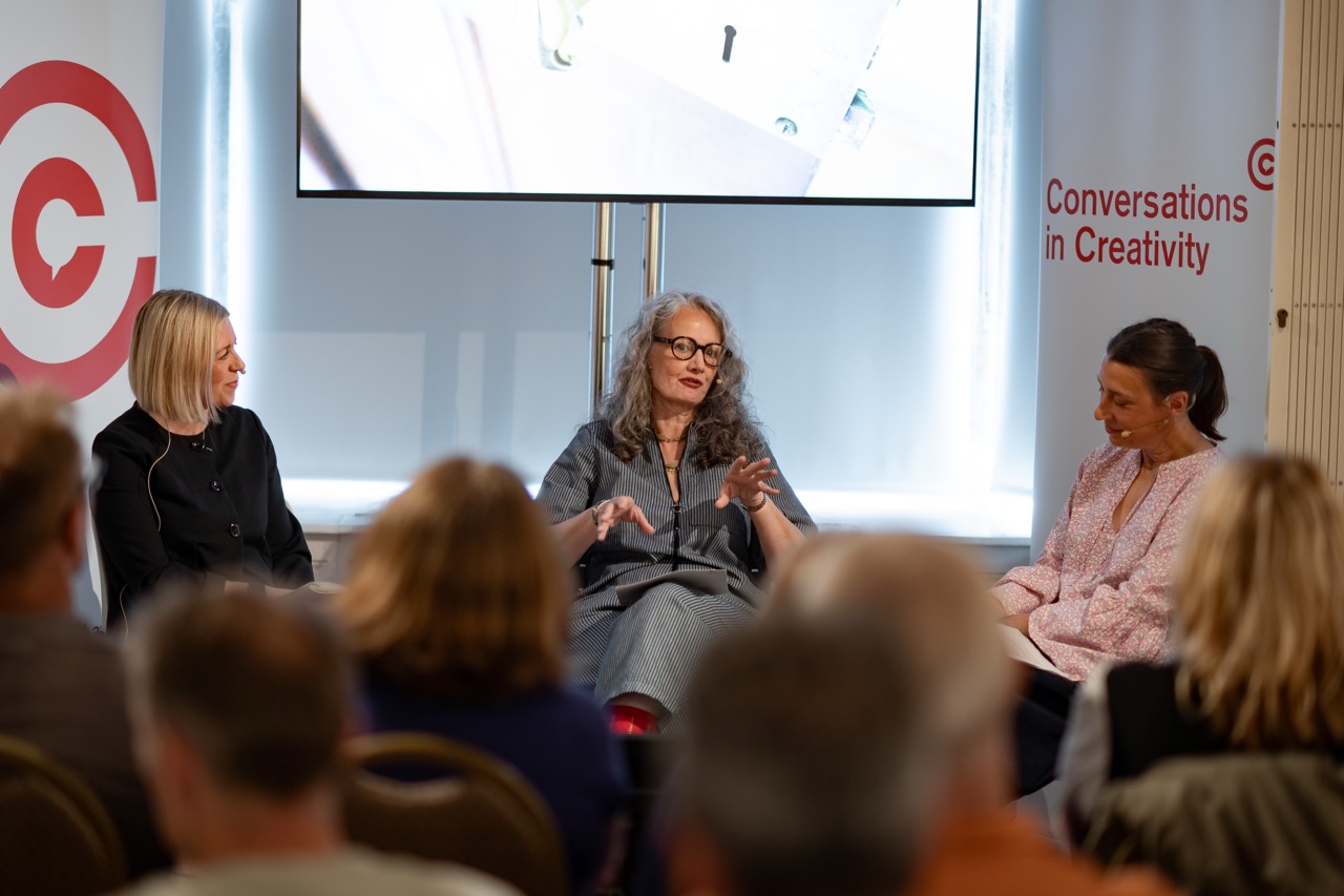 Three women sit on stage in conversation during an event titled Conversations in Creativity, speaking to an attentive audience. A large screen and branded banners are visible behind them.
