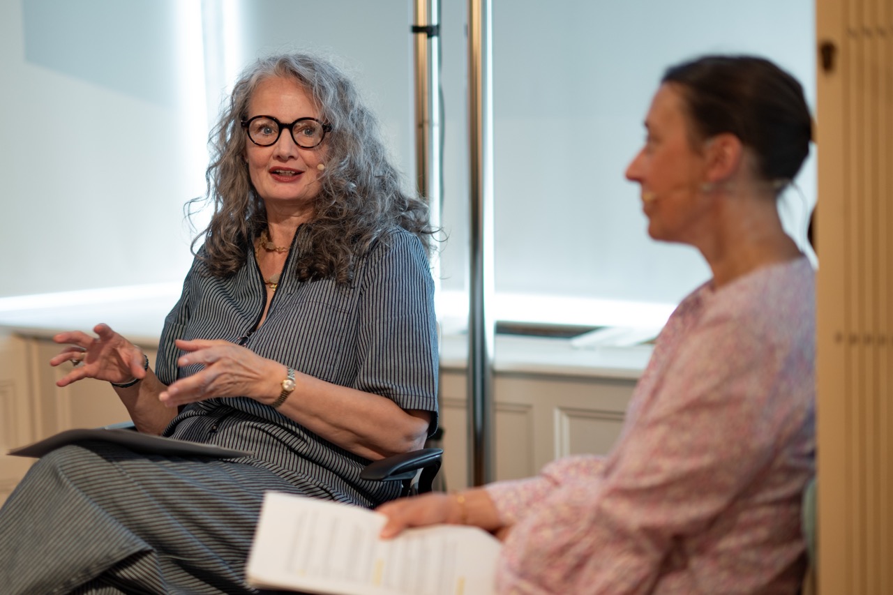 Two women sit indoors having a conversation. One gestures with her hands while speaking, holding papers in her lap. Both wear microphones and casual clothing, with soft lighting in the background.