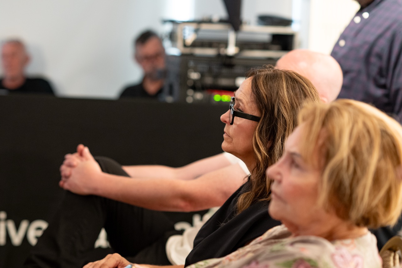 Three adults sit closely together in an indoor setting, attentively facing the same direction. The woman in the centre wears glasses and looks focused. Other people and equipment are visible in the blurred background.
