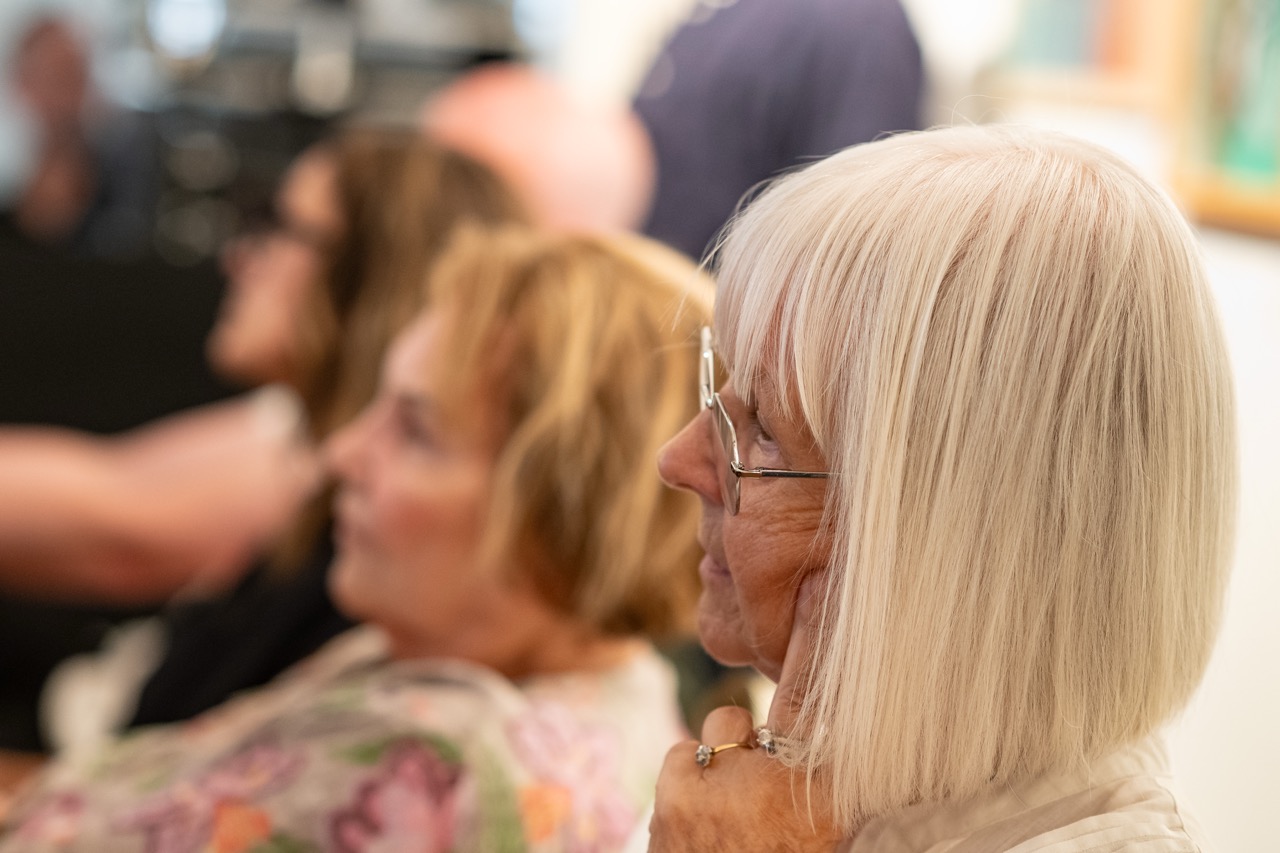 An older woman with white hair and spectacles sits in focus, listening intently. Other people are blurred in the background, suggesting a group setting or audience.