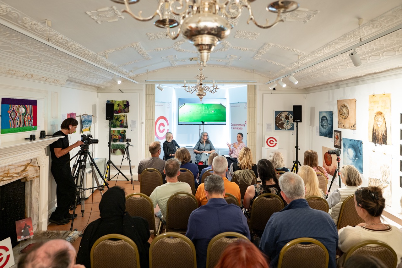 An audience watches a panel discussion in an art gallery. Three speakers sit at the front, whilst a cameraman records. Artworks are displayed on the walls, and a chandelier hangs from an ornate ceiling.