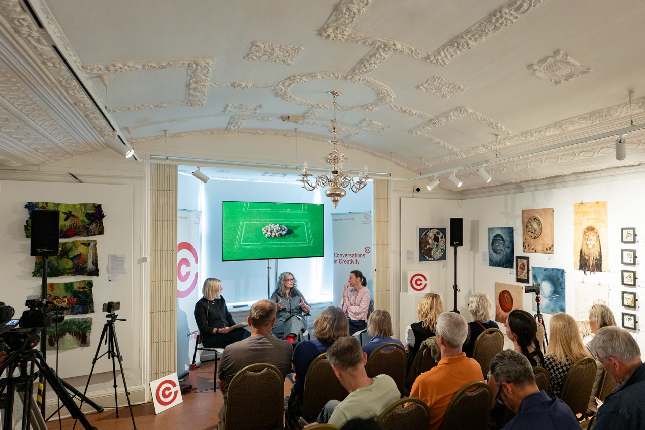 Three people sit onstage in front of an audience for a panel discussion in an art gallery. Behind them, art is displayed on the walls and a large screen shows an image of sheep on green grass.