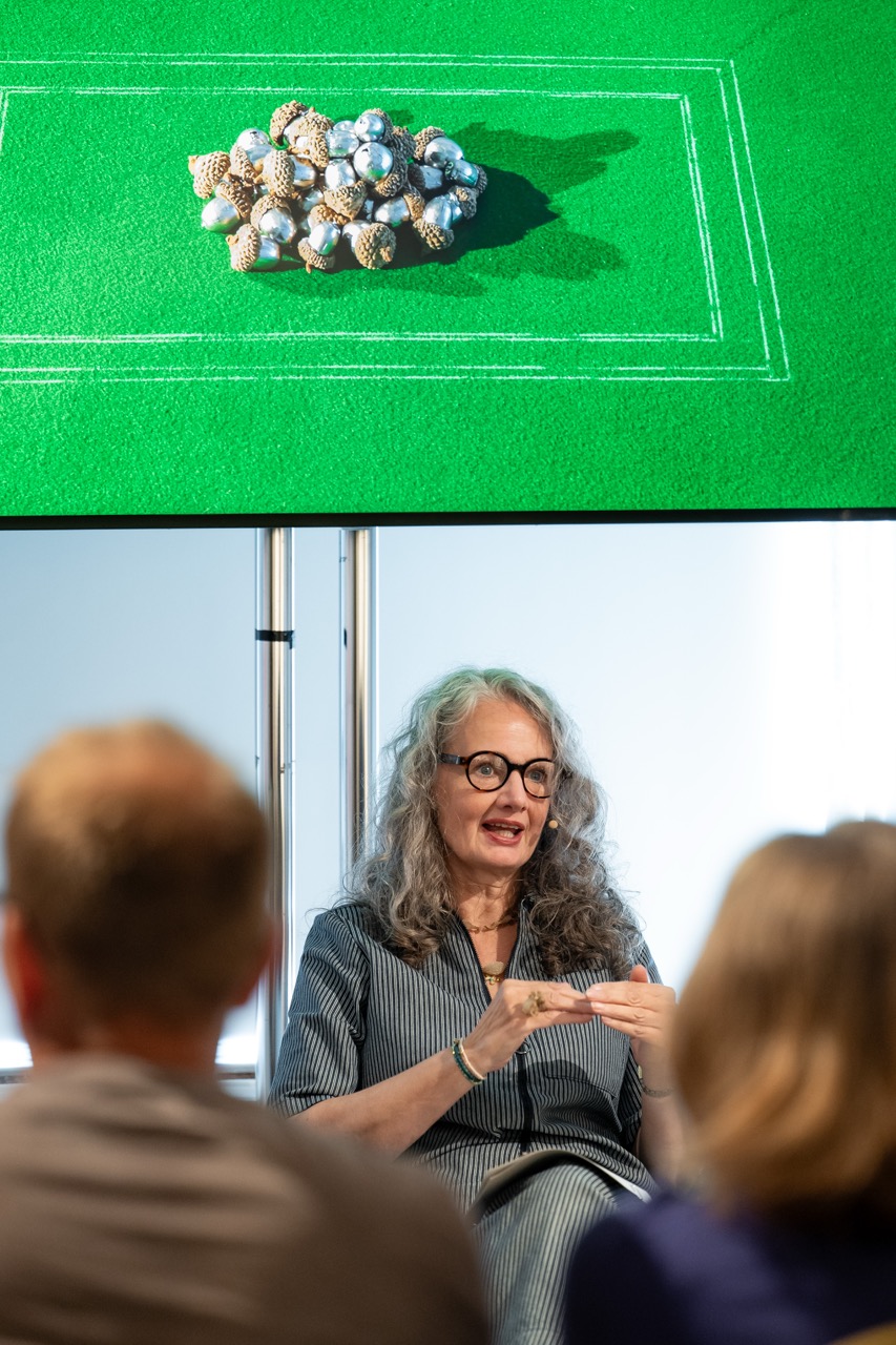 A woman with grey hair and glasses speaks to an audience. Above her is a green screen displaying a cluster of spheres and cones, outlined by white lines.