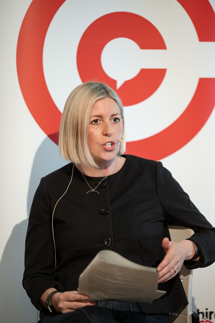 A woman with short blonde hair speaks while holding papers, wearing a black top and headset mic, seated in front of a large red and white circular logo.