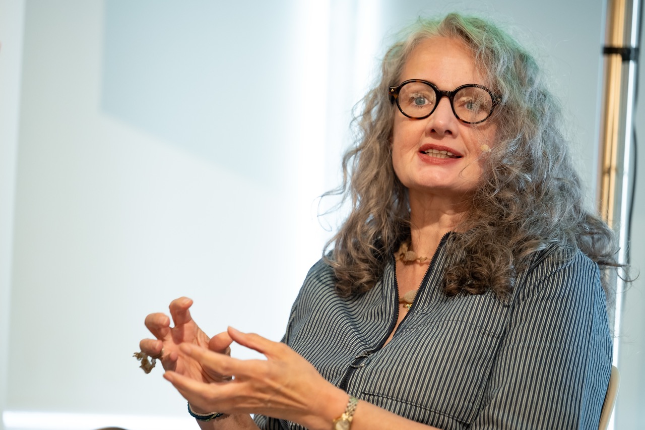 A woman with long curly grey hair and glasses gestures with her hands while speaking, wearing a striped shirt and a necklace, with a blurred light background.