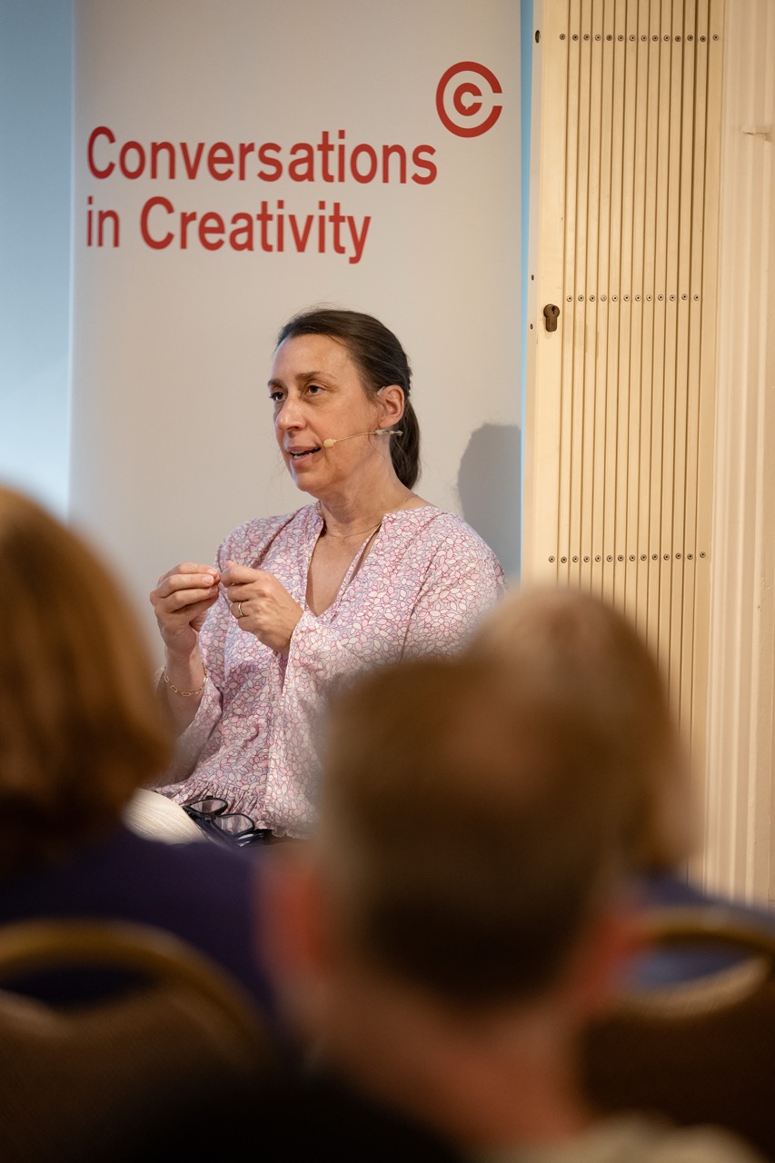 A woman speaks to an audience in front of a sign that reads Conversations in Creativity. She gestures with her hands whilst engaged in discussion.