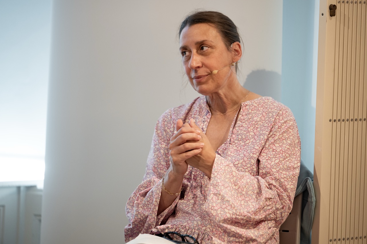 A woman with a headset microphone sits, clasping her hands, wearing a light pink patterned blouse, looking slightly to the side in a well-lit room.