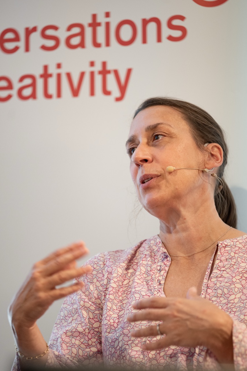 A woman wearing a pale patterned blouse speaks into a headset microphone, gesturing with her hands. The background shows partially visible red text reading Conversations Creativity.