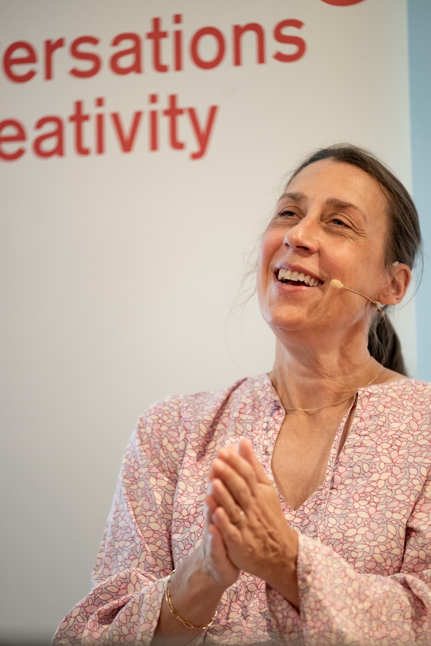 A woman wearing a light patterned blouse and headset microphone smiles and clasps her hands. Behind her, Conversations and Creativity are partially visible on a white background.