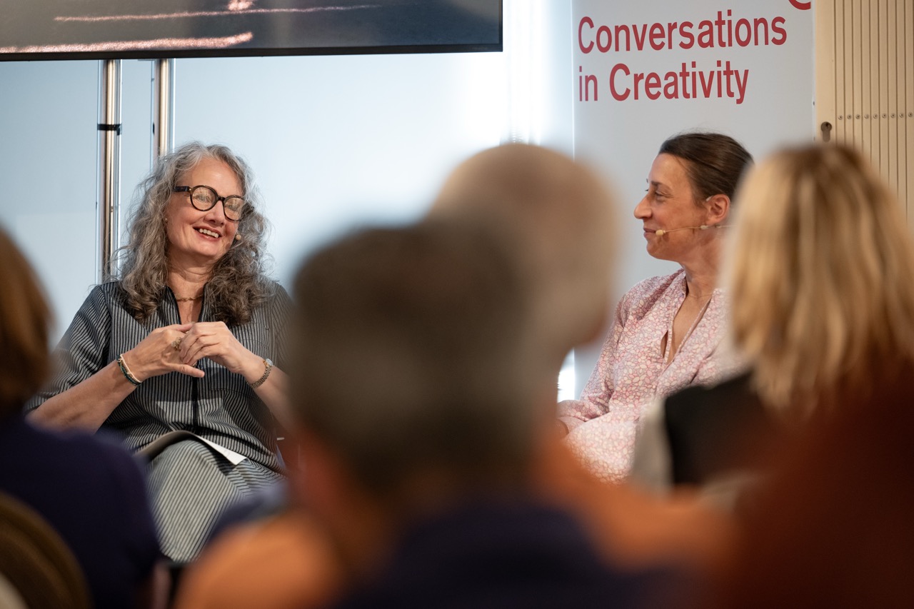 Two women are seated and speaking at an event. One is smiling and gesturing with her hands, whilst the other listens attentively. A banner behind them reads Conversations in Creativity. Audience members are visible in the foreground.