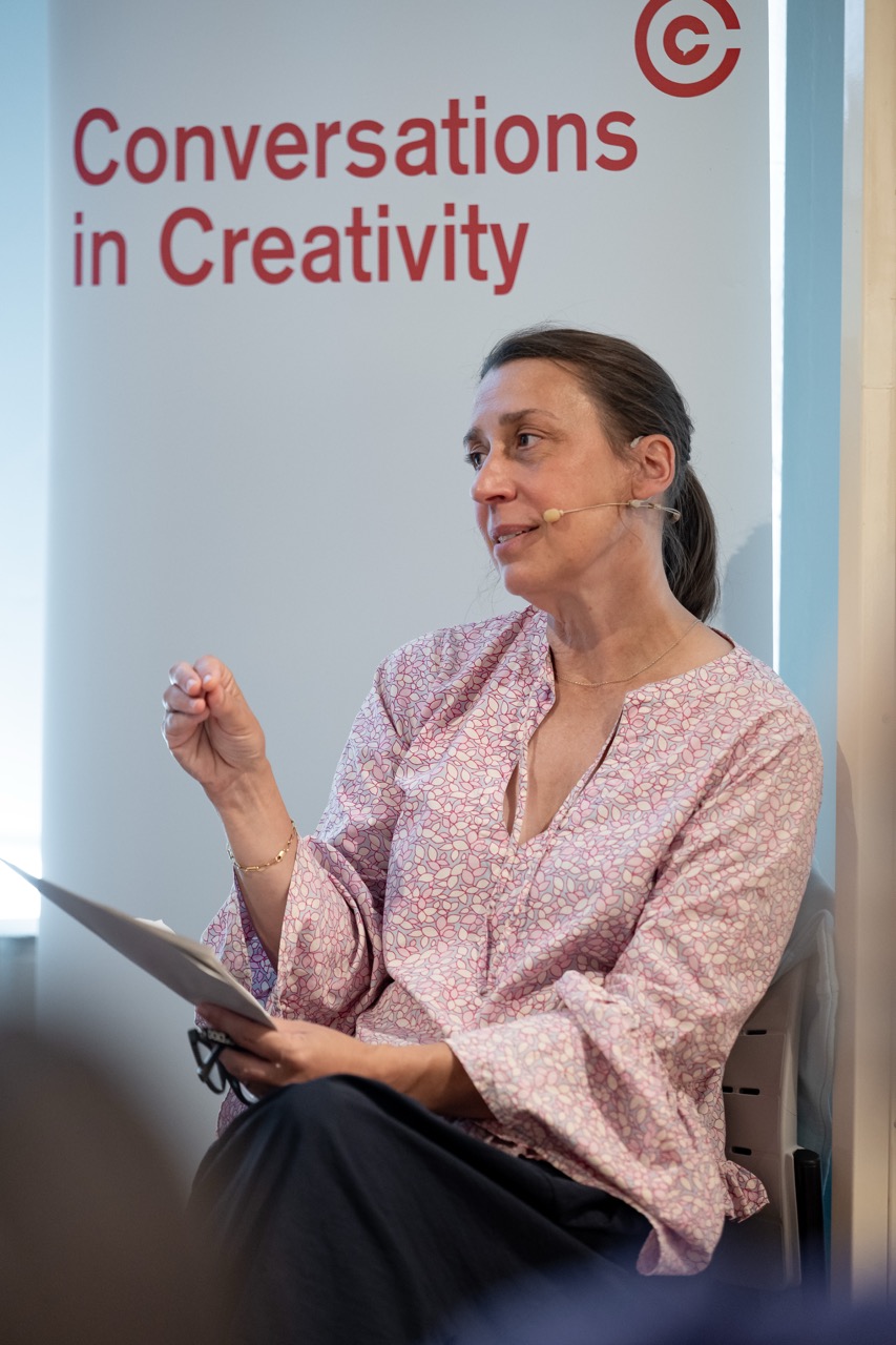 A woman with a headset microphone speaks while holding papers, seated in front of a banner that reads Conversations in Creativity.