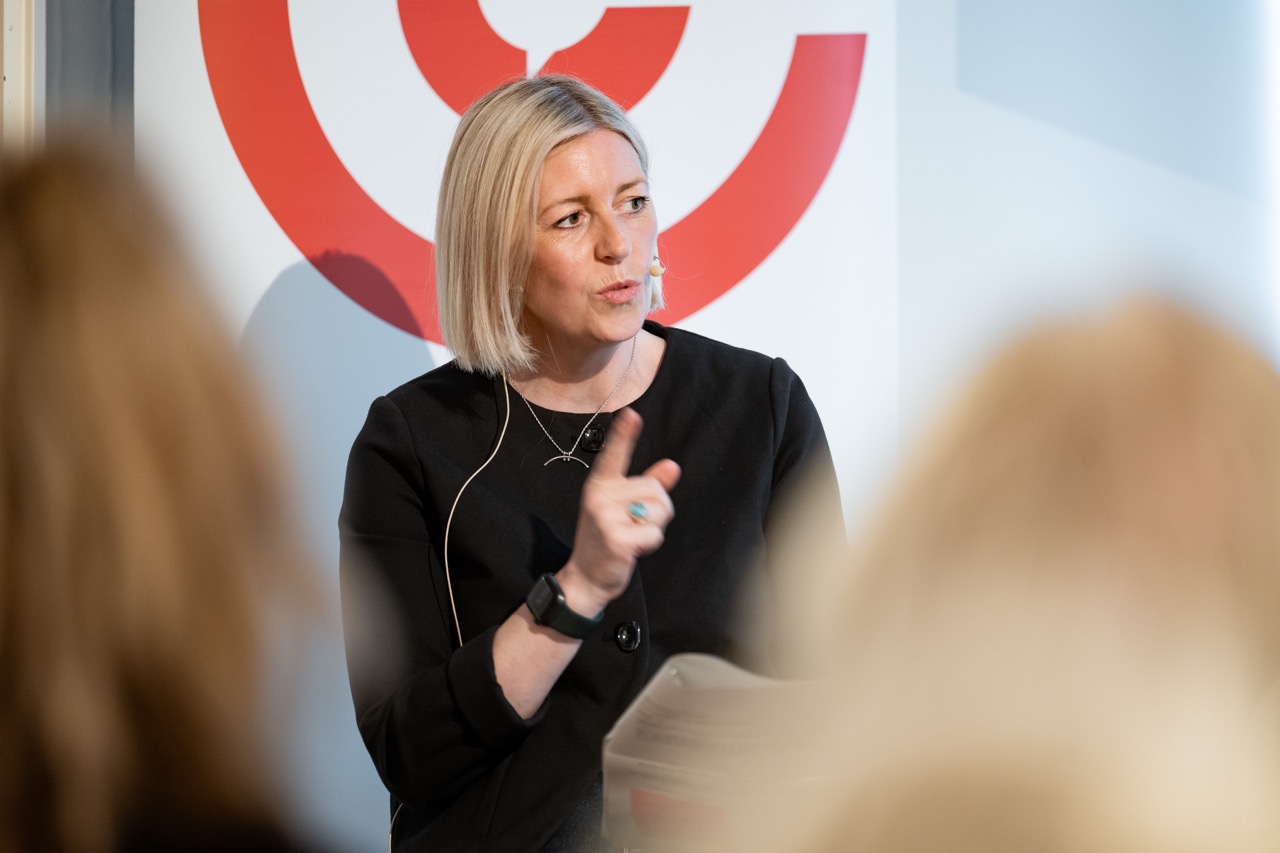 A woman with blonde hair speaks whilst gesturing with her hand. She wears a black outfit and a headset microphone, sitting in front of a red and white circular design. Blurred audience members are visible in the foreground.