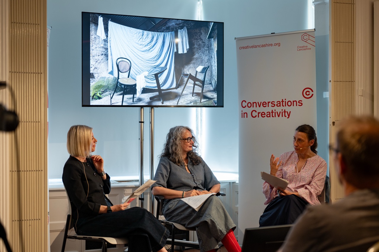Three women sit and talk in front of a TV displaying an artistic photo. A banner reads Conversations in Creativity. The setting appears to be an indoor creative discussion or interview event.